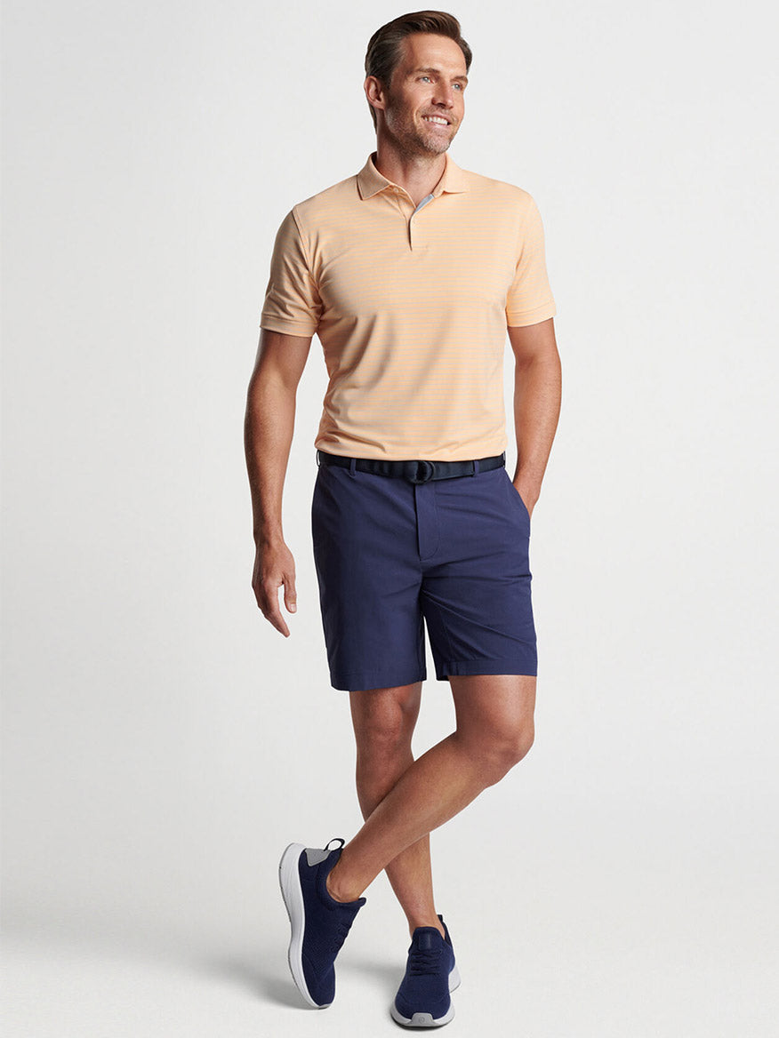 Man posing in a casual tan polo shirt and Peter Millar Surge Performance Short in Navy with matching sneakers, featuring four-way stretch, water-resistant performance apparel.