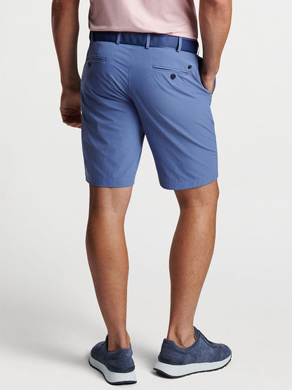 Man wearing Peter Millar Surge Performance Short in Blue Pearl with four-way stretch and sneakers, standing with hands in pockets.