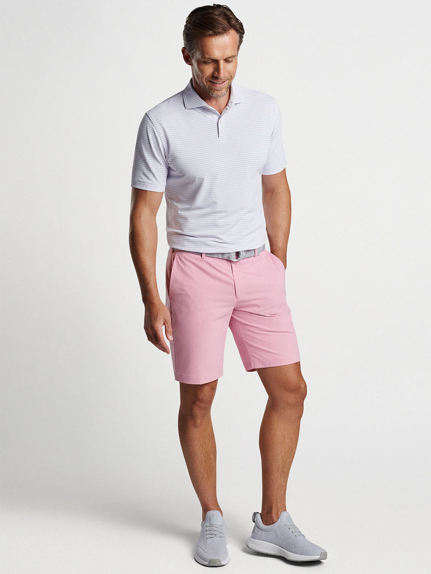 A man wearing a light striped polo shirt featuring four-way stretch, Peter Millar Surge Performance Shorts in Spring Blossom with water-resistant protection, and gray sneakers poses against a neutral background.