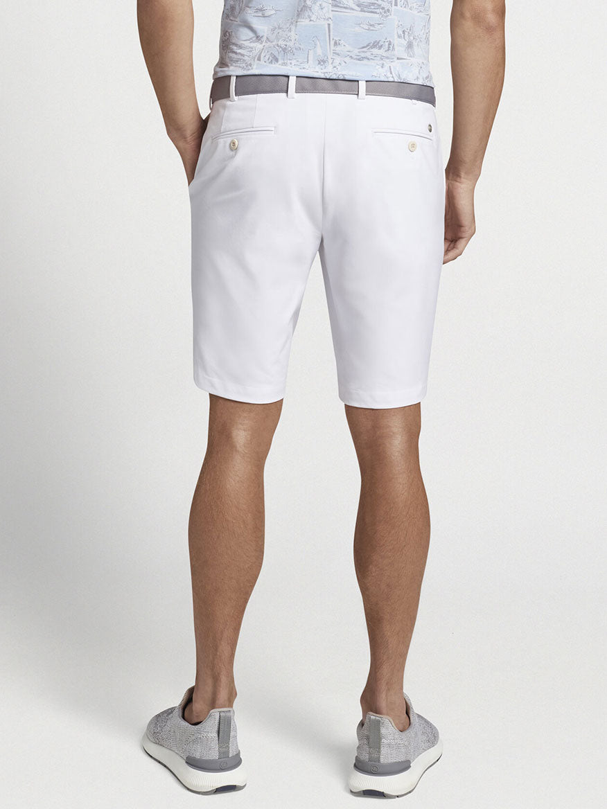 Man wearing Peter Millar Surge Performance Shorts in White with water-resistant protection, grey sneakers, and a patterned performance apparel shirt viewed from behind.