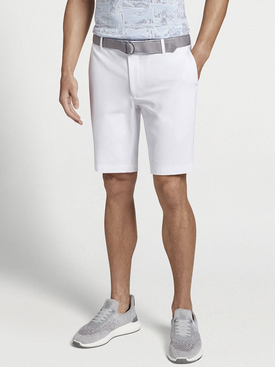 A man stands showcasing the Peter Millar Surge Performance Short in White with wicking ability, gray sneakers, and a light belt, with the upper body not visible.