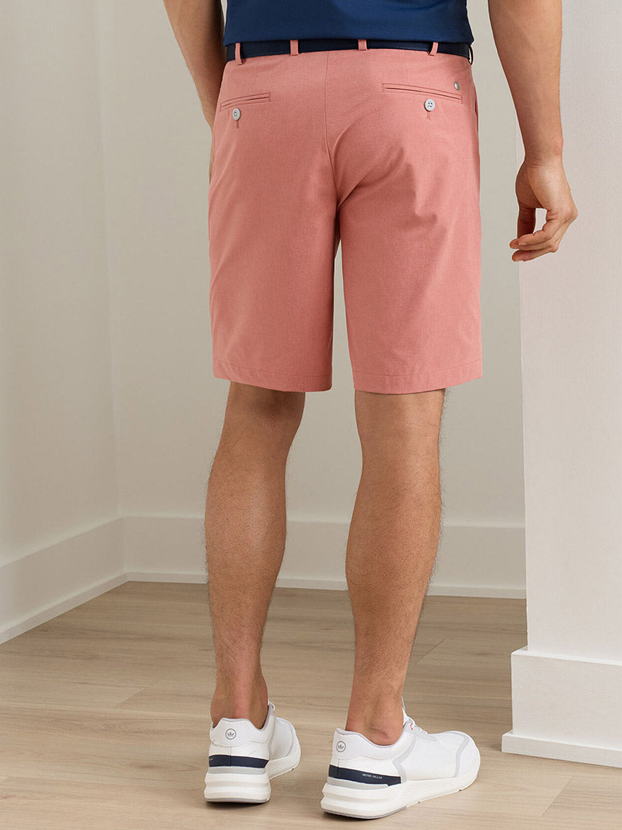 A person wearing Peter Millar Surge Performance Short in Faded Scarlet, a navy shirt, and white sneakers stands indoors facing away from the camera.