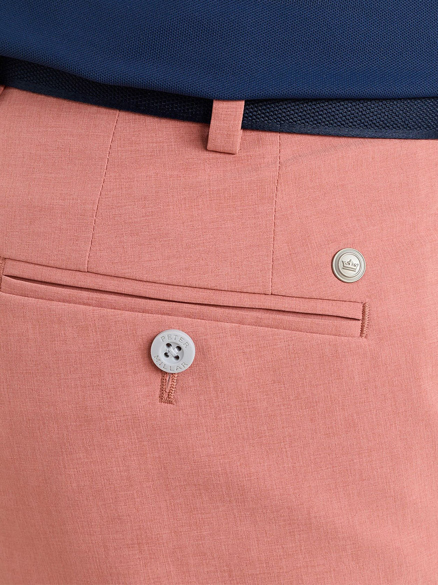 Close-up of the back pocket area of Peter Millar Surge Performance Short in Faded Scarlet, featuring a small button and metal emblem, styled with a navy blue belt and shirt.