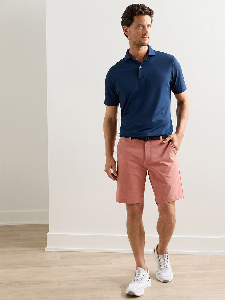 A man wearing a navy polo, Peter Millar Surge Performance Short in Faded Scarlet, white sneakers, and a belt stands indoors on a light wood floor against a plain wall.