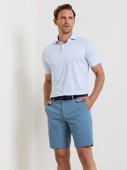 A man stands against a plain wall wearing a light blue polo shirt tucked into Peter Millar Surge Performance Short in Storm, styled with a black belt, hands in his pockets, and looking slightly to the side.
