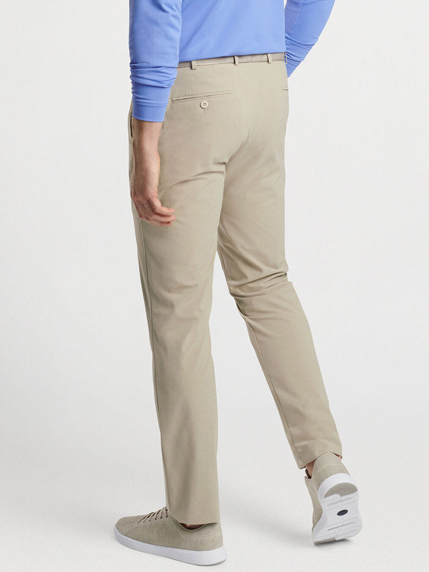 A person wearing a light blue, long-sleeve shirt made of four-way stretch material, Peter Millar Surge Performance Trouser in Toffee, and light gray sneakers seen from the back.