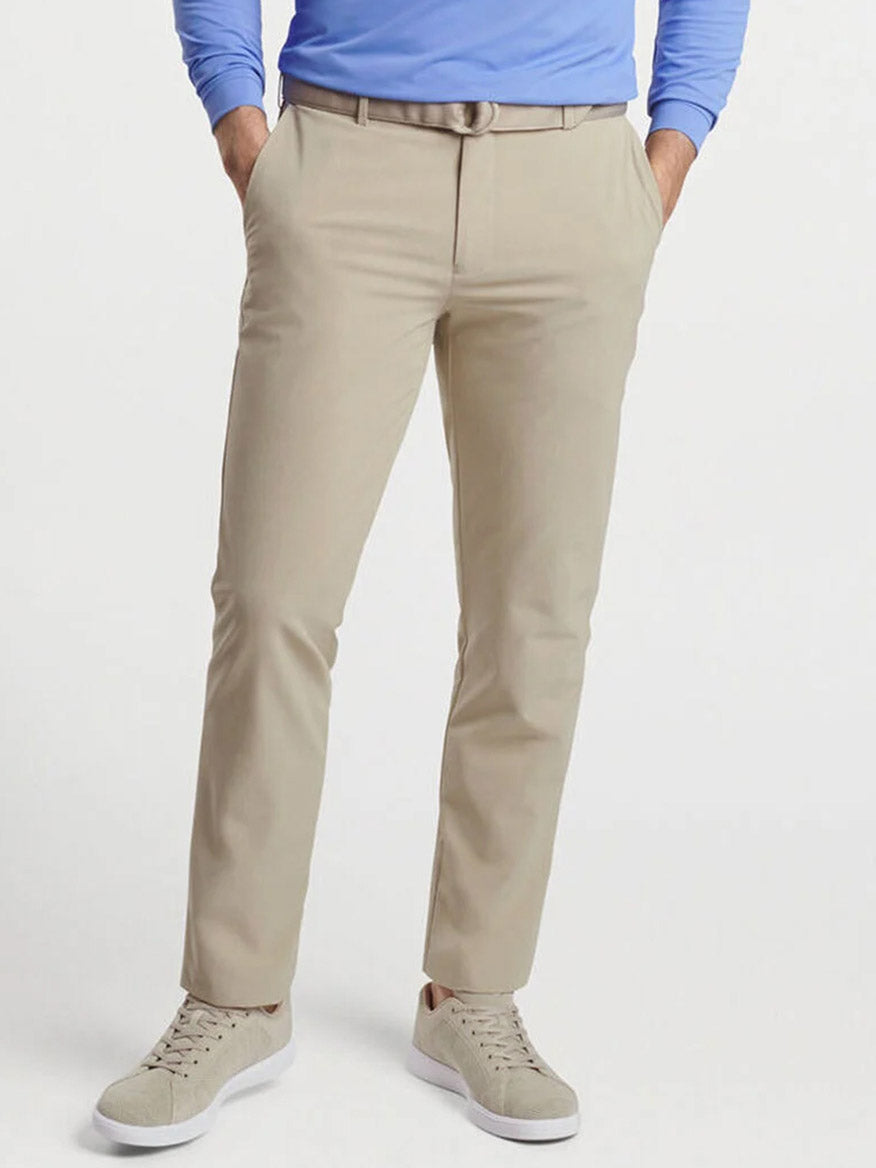 Person wearing Peter Millar Surge Performance Trouser in Toffee with pockets made from four-way stretch fabric, a light blue long-sleeve shirt, tan belt, and beige sneakers, standing against a plain white background.