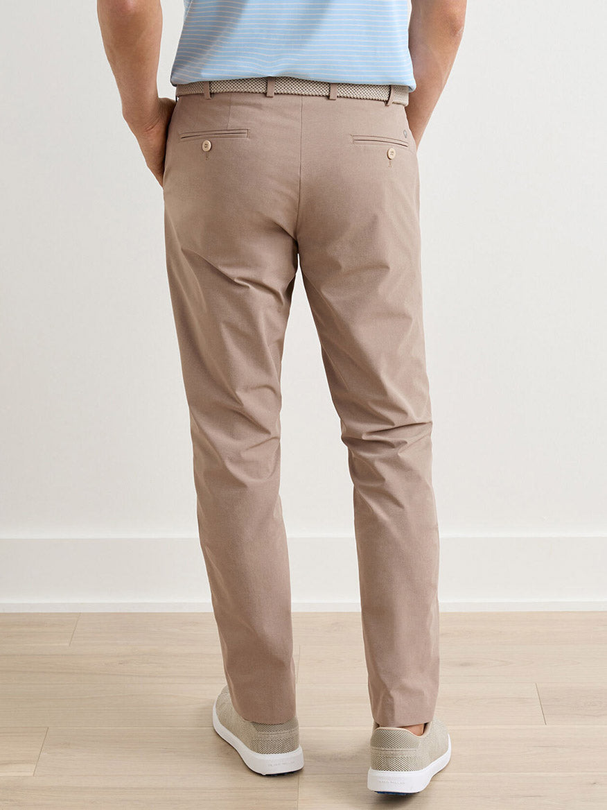 A person stands indoors, seen from the back, wearing Peter Millar Surge Performance Trouser in Biscotti with four-way stretch, a light blue shirt, a beige belt, and light-colored slip-on shoes.