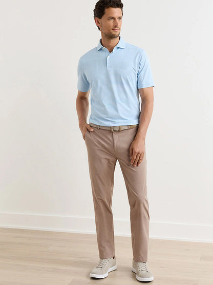 A man stands indoors wearing a light blue polo shirt, Peter Millar Surge Performance Trouser in Biscotti with four-way stretch, a matching belt, and light gray sneakers, his left hand in his pocket.