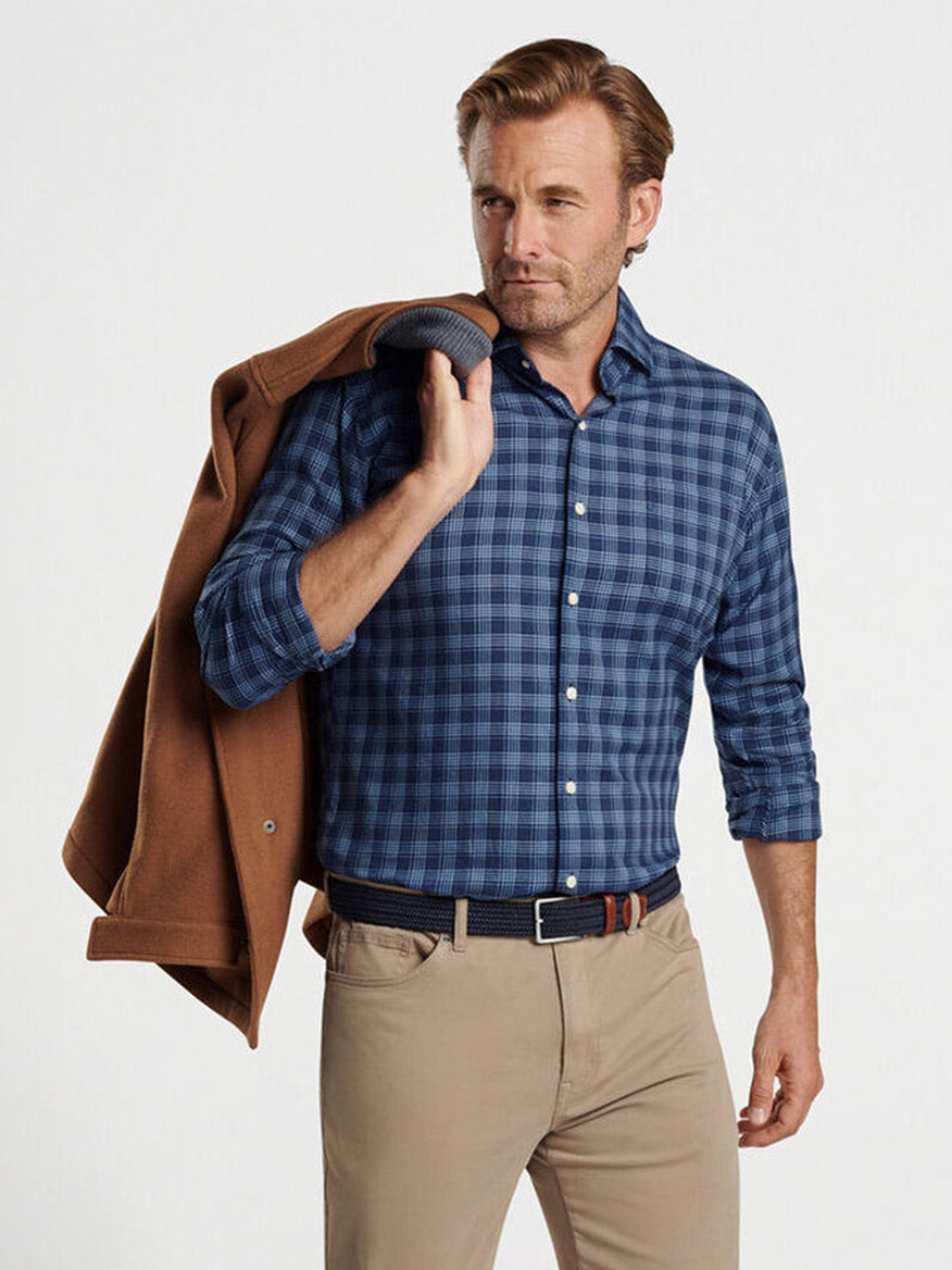 Wearing the Peter Millar Thompson Summer Soft Cotton Sport Shirt in Galaxy, a man stylishly drapes a brown jacket over his shoulder. Set against a plain background, he complements the classic fit blue plaid shirt with beige pants and a dark belt, embodying effortless style.