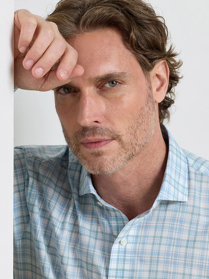 A man with light brown hair and a trimmed beard wears the Peter Millar Tropea Italian Poplin Sport Shirt in Light Willow, resting his hand on his forehead and looking at the camera against a plain background.