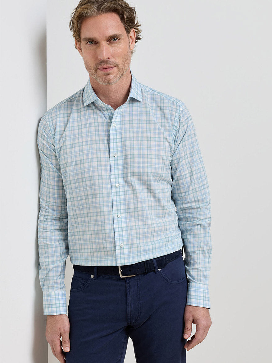 Man standing against a white wall, wearing the Peter Millar Tropea Italian Poplin Sport Shirt in Light Willow, tailored fit, paired with dark blue pants and a black belt.