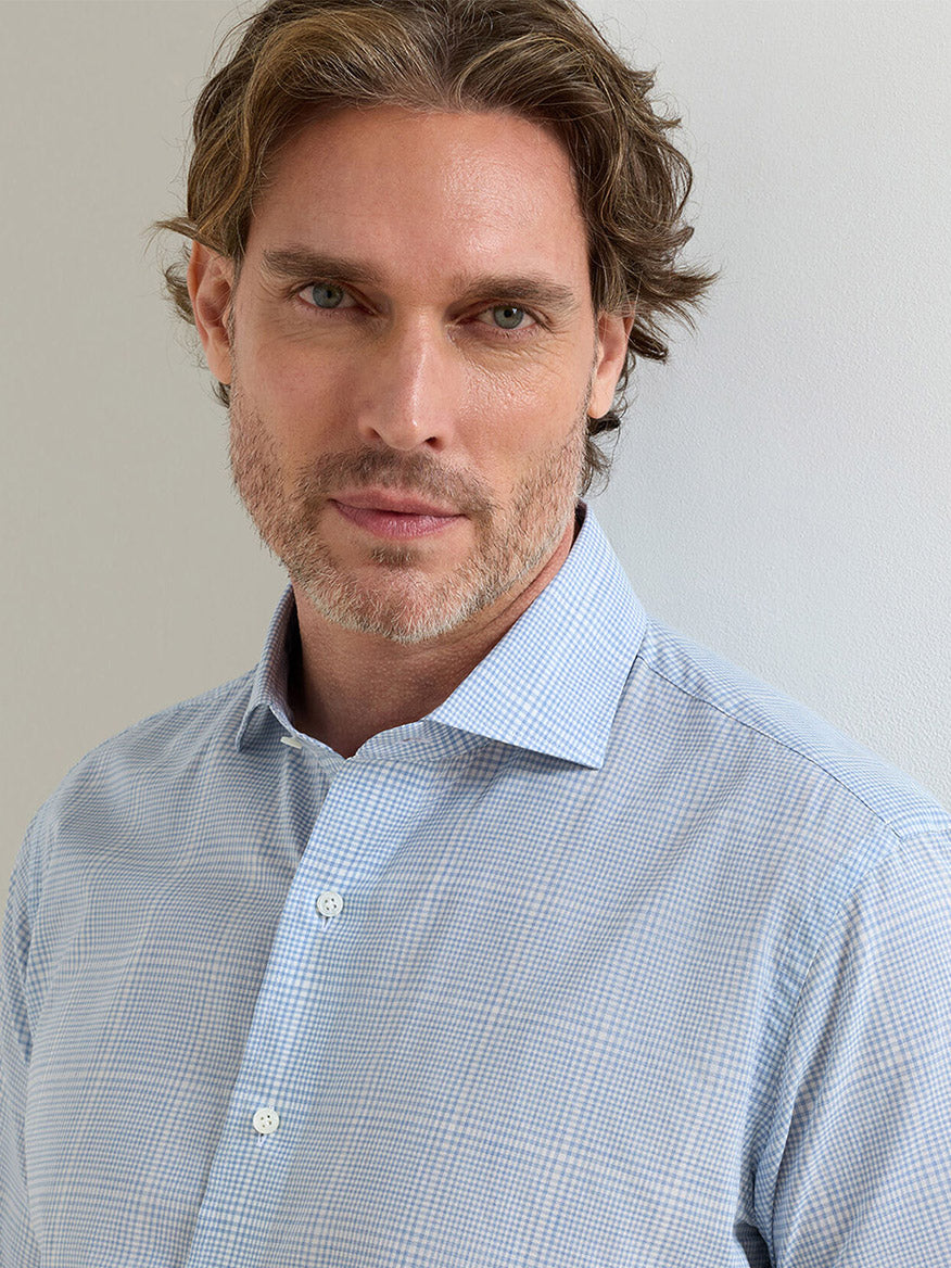 A man with light brown hair and a short beard wears the Peter Millar Urbino Italian Poplin Sport Shirt in Iris Mist, featuring a tailored fit and light blue check pattern, standing against a plain light-colored wall.