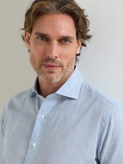 A man with light brown hair and a short beard wears the Peter Millar Urbino Italian Poplin Sport Shirt in Iris Mist, featuring a tailored fit and light blue check pattern, standing against a plain light-colored wall.