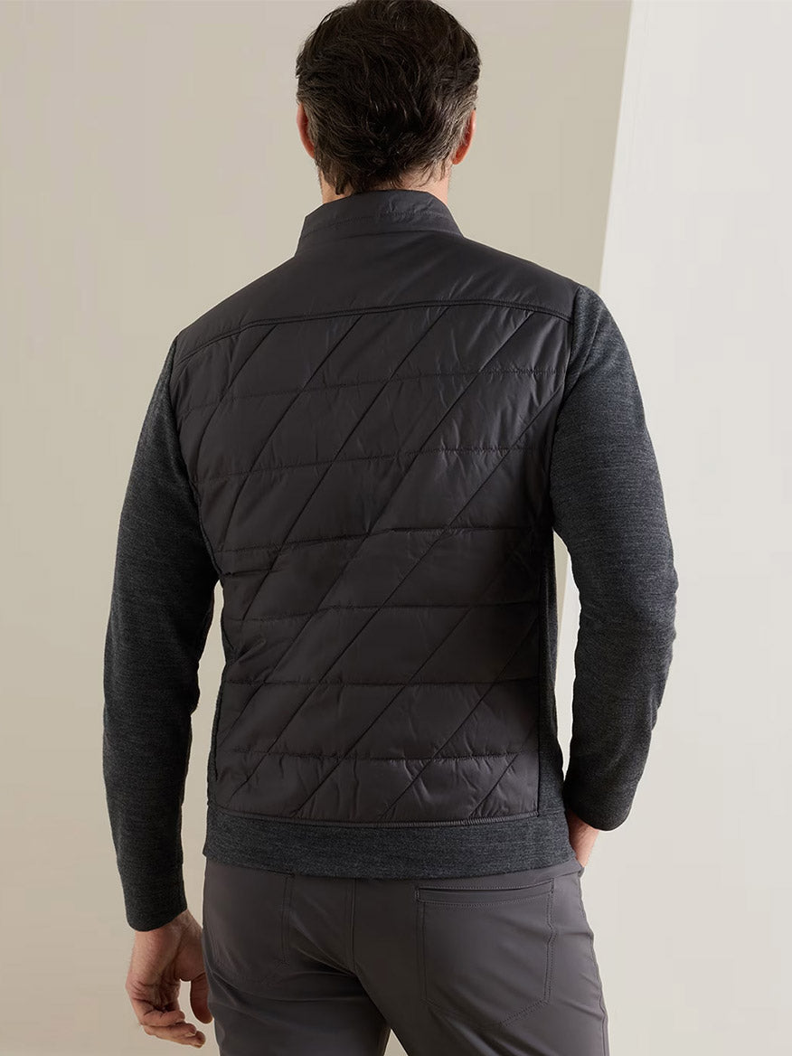 A man faces away from the camera, wearing the Peter Millar Walker Hybrid Jacket in Iridium—a quilted, water-resistant jacket with textured sleeves—paired with matching gray pants against a light-colored wall.
