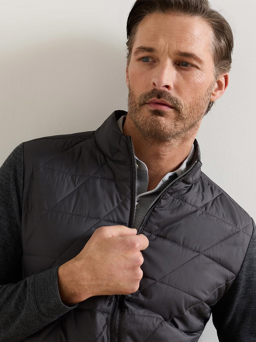 A man with light brown hair and stubble stands against a light-colored wall, looking slightly to the side while holding the zipper of his Peter Millar Walker Hybrid Jacket in Iridium, layered over a gray shirt.