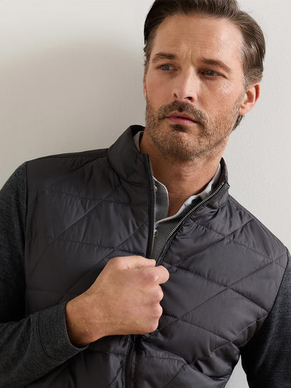 A man with light brown hair and stubble stands against a light-colored wall, looking slightly to the side while holding the zipper of his Peter Millar Walker Hybrid Jacket in Iridium, layered over a gray shirt.