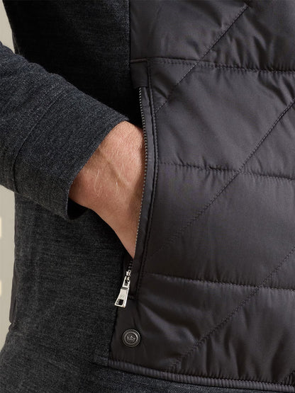 A person wearing the Peter Millar Walker Hybrid Jacket in Iridium, a dark quilted jacket featuring both a zipper and snap buttons, with their left hand in a side pocket.