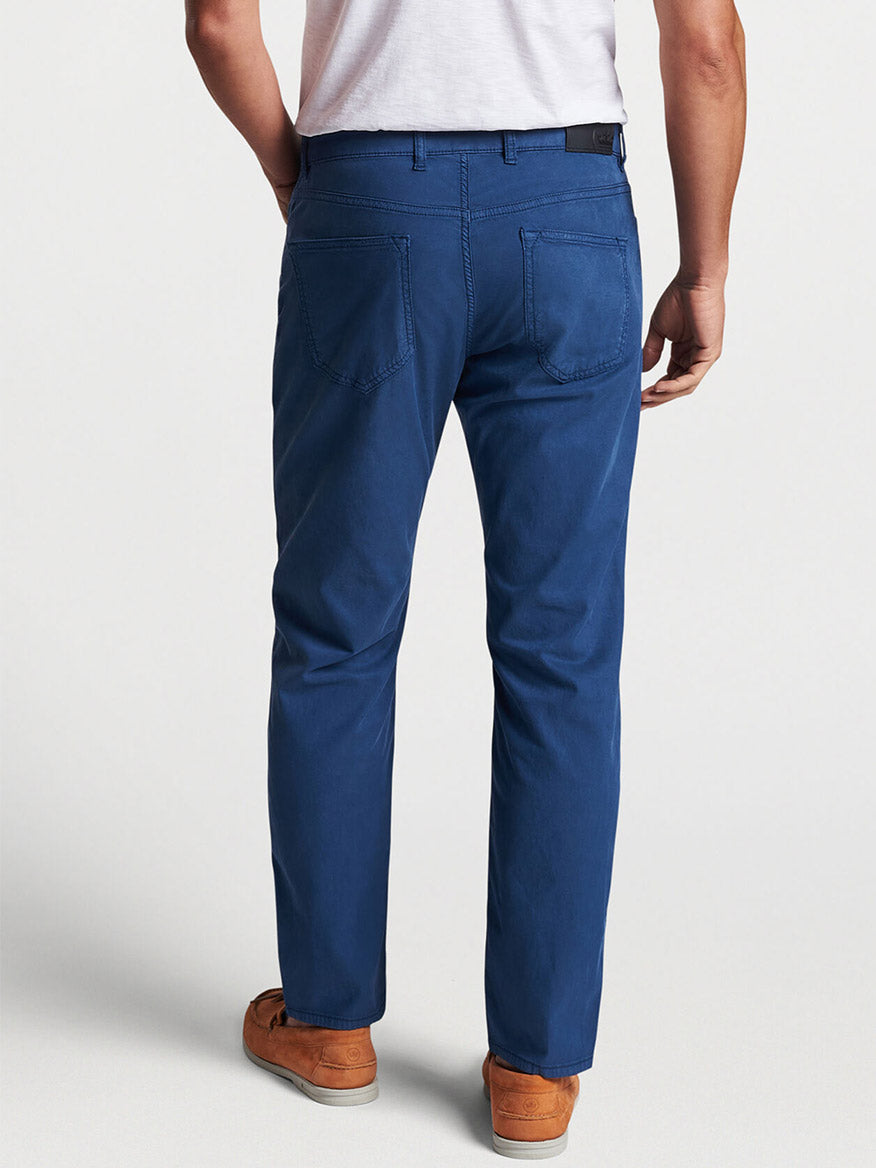 Rear view of a person wearing blue jeans and brown shoes standing with one hand in a Peter Millar Wayfare Five-Pocket Pant in Riviera Blue pocket.