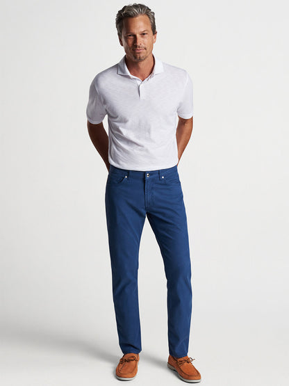 Man in a white polo shirt and Peter Millar Wayfare Five-Pocket Pant in Riviera Blue, tailored for a comfortable stretch, posing with hands in pockets.