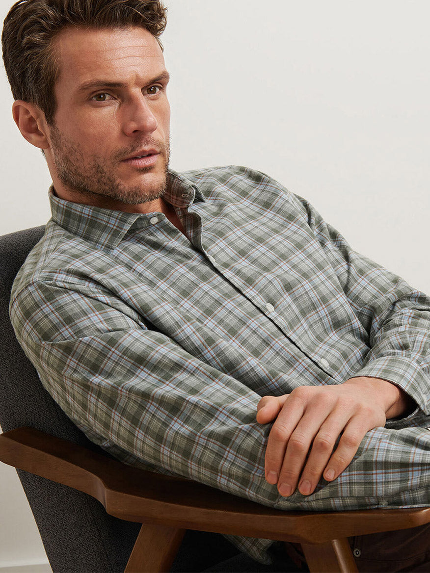 A man with short brown hair and facial stubble sits in a chair, wearing a Peter Millar Whitecap Performance Flannel Sport Shirt in Basil.
