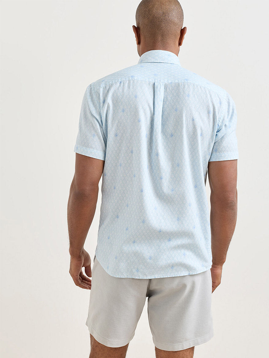 A man stands facing away, wearing a Peter Millar Whitehaven Cotton Lyocell Sport Shirt in Stream Blue and light gray shorts against a plain background.