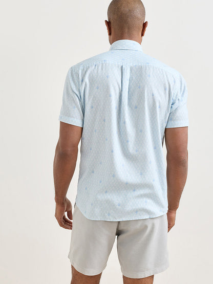 A man stands facing away, wearing a Peter Millar Whitehaven Cotton Lyocell Sport Shirt in Stream Blue and light gray shorts against a plain background.