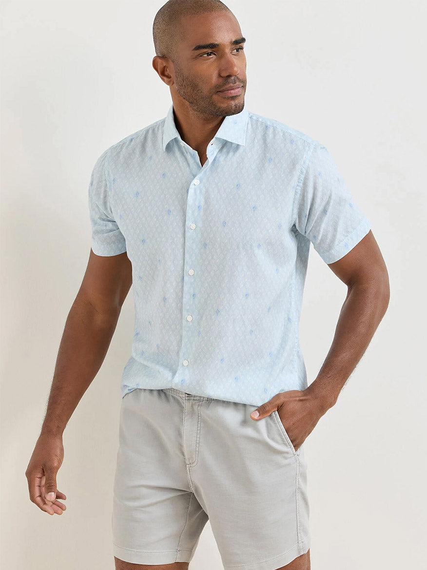 A man wearing a Peter Millar Whitehaven Cotton Lyocell Sport Shirt in Stream Blue and light gray shorts stands with one hand in his pocket against a plain background.