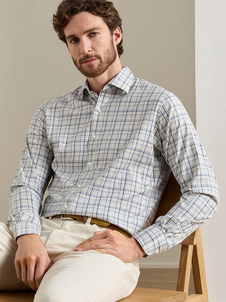 A man with a beard and wavy hair sits on a wooden chair, wearing the Peter Millar Wyatt Crown Soft Twill Sport Shirt in Multi—crafted from easy-care, stretch performance fabric—and cream-colored pants.