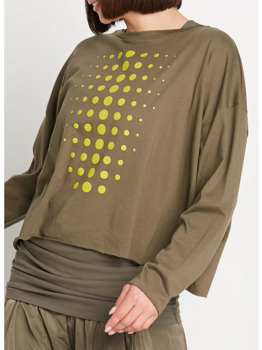 A person is wearing the Planet by Lauren G Dot Dot Dot Mini Luxury Boxy Tee in Loden/Guacamole, a loose-fitting, long-sleeved olive green top featuring playful dots arranged in a triangular pattern. Their lower garment is partially visible.