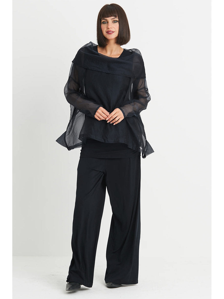 Against a plain background, the person stands in the Planet by Lauren G Organza Cowl Neck Top in Black layered over loose dark trousers, adding a subtle feminine touch.