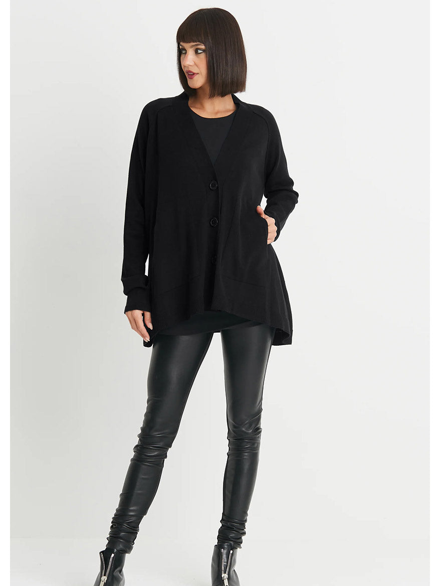 A person with chin-length dark hair, wearing the Planet by Lauren G Varsity Cardigan in black, along with a black top, black leather pants, and black ankle boots, stands against a plain white background.