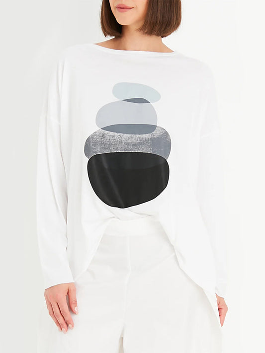 A person wearing a white Planet by Lauren G Zen Luxury Boxy T with an abstract graphic of stacked oval shapes in varying shades of blue and black. The shirt, Zen-inspired in print, is paired with white pants. The face is not visible.