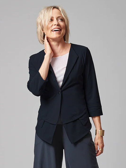 A smiling woman with short blonde hair, wearing a Porto Bonton Jacket in Shadow, white top, and dark trousers, standing with one hand on her neck against a gray background.