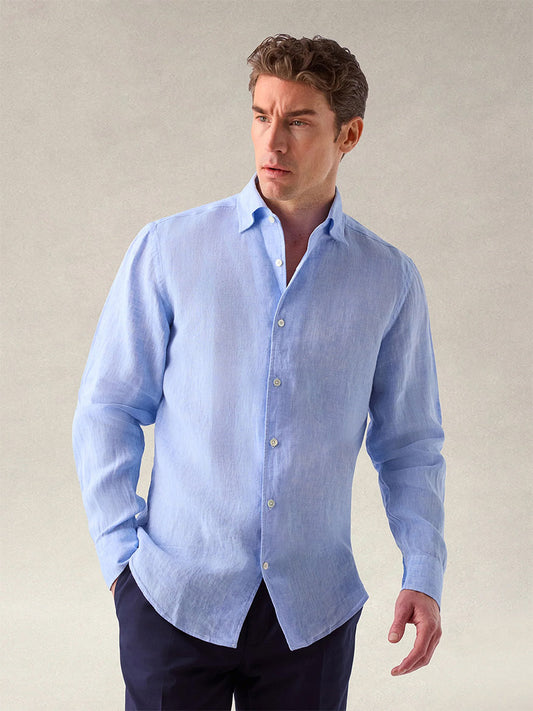 A man wears the Robert Talbott Morgan Linen Shirt in Blue, featuring breathable, lightweight French linen and a one-piece collar, paired with dark pants as he stands against a plain, light background, looking slightly to the side.