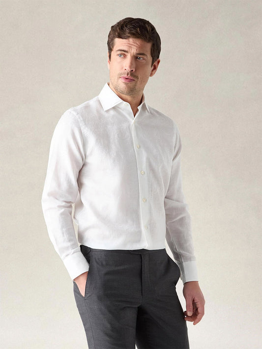 A man stands against a plain light background, one hand in his pocket, wearing the Robert Talbott Reynolds Paisley Jacquard Dress Shirt in White with tailored construction and dark pants.
