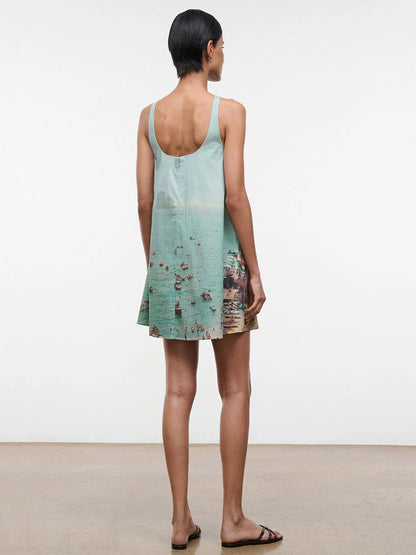 A woman stands indoors facing away, wearing the STAUD Brio Dress in Endless Summer with a beach and water print—perfect for warm weather. She pairs it with black sandals; her short dark hair is seen above a light brown floor.