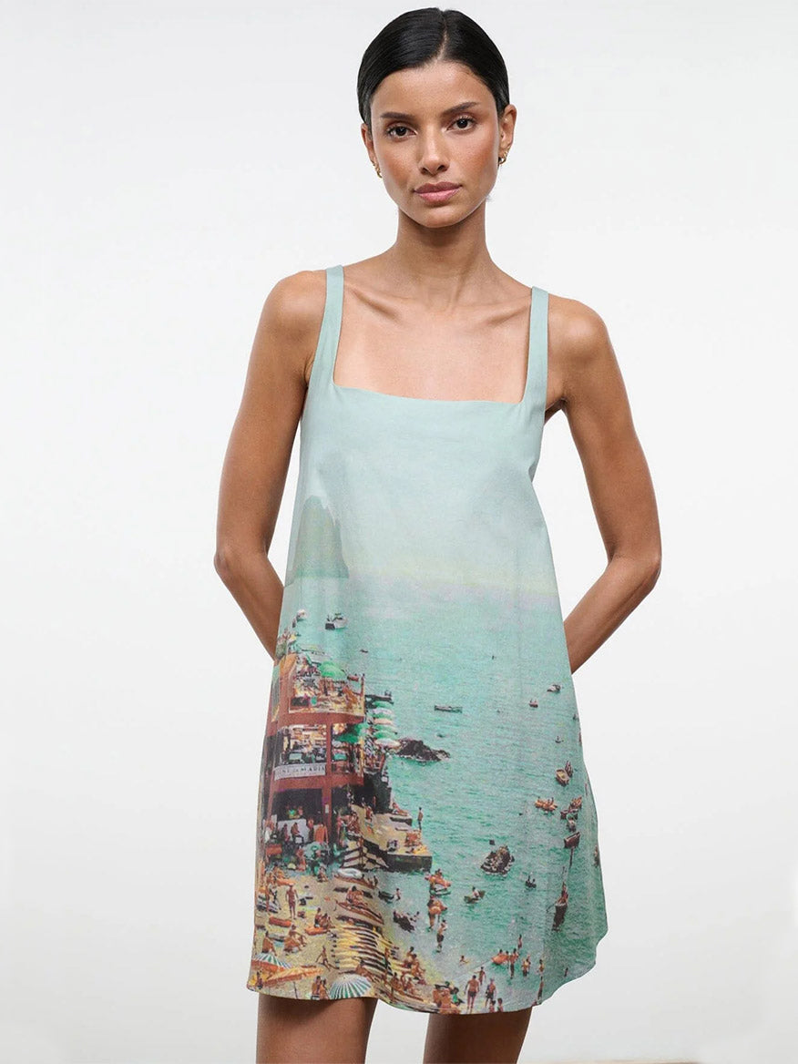 A woman stands against a plain background wearing the STAUD Brio Dress in Endless Summer, a sleeveless pull-on tent dress featuring a seaside print—ideal for effortless warm-weather style.