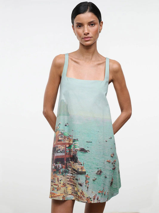 A woman stands against a plain background wearing the STAUD Brio Dress in Endless Summer, a sleeveless pull-on tent dress featuring a seaside print—ideal for effortless warm-weather style.