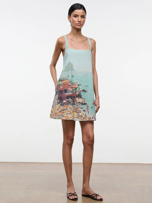 A woman stands against a plain background wearing the STAUD Brio Dress in Endless Summer—a sleeveless, pull-on tent silhouette with a printed seaside scene—styled with black sandals for effortless warm-weather dressing.