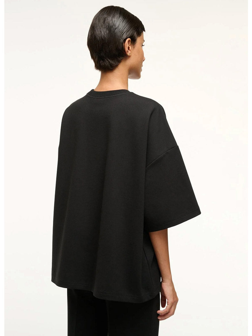 A person with short dark hair is seen from the back, wearing the STAUD Capsule Top in Black—an oversized crewneck shirt styled with black ponte pants—perfect as a travel day essential, set against a plain light background.