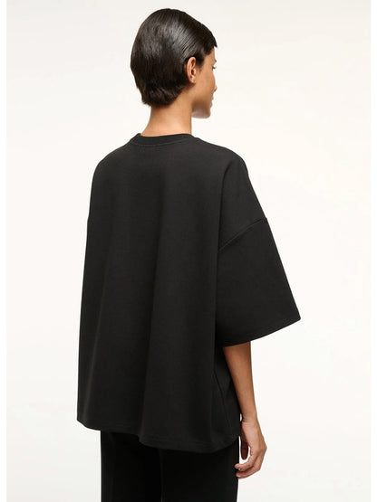 A person with short dark hair is seen from the back, wearing the STAUD Capsule Top in Black—an oversized crewneck shirt styled with black ponte pants—perfect as a travel day essential, set against a plain light background.