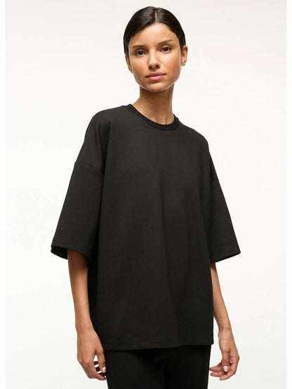 A person stands facing forward, wearing the STAUD Capsule Top in Black—an oversized crewneck—paired with black pants, travel day essentials, against a plain light background.