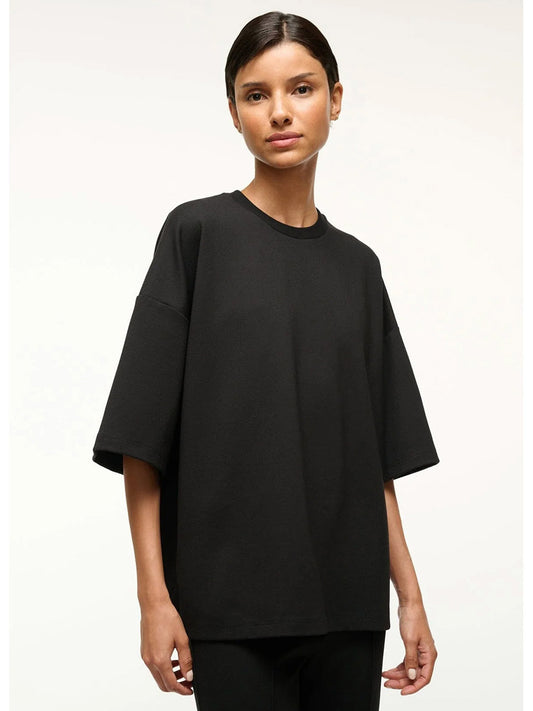 A person stands facing forward, wearing the STAUD Capsule Top in Black—an oversized crewneck—paired with black pants, travel day essentials, against a plain light background.