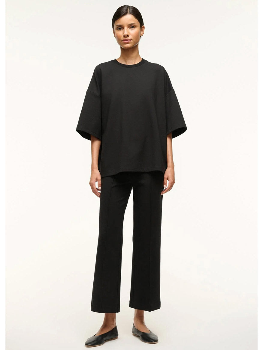 A woman faces forward, wearing the STAUD Capsule Top in Black—an oversized crewneck shirt—paired with black ponte straight-leg pants, ideal for travel days, set against a plain light background.