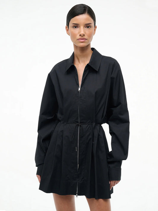 A person faces forward against a white background wearing the STAUD Chi Dress in Black, featuring long sleeves, stretch poplin fabric, a zippered front, and an adjustable waist tie.