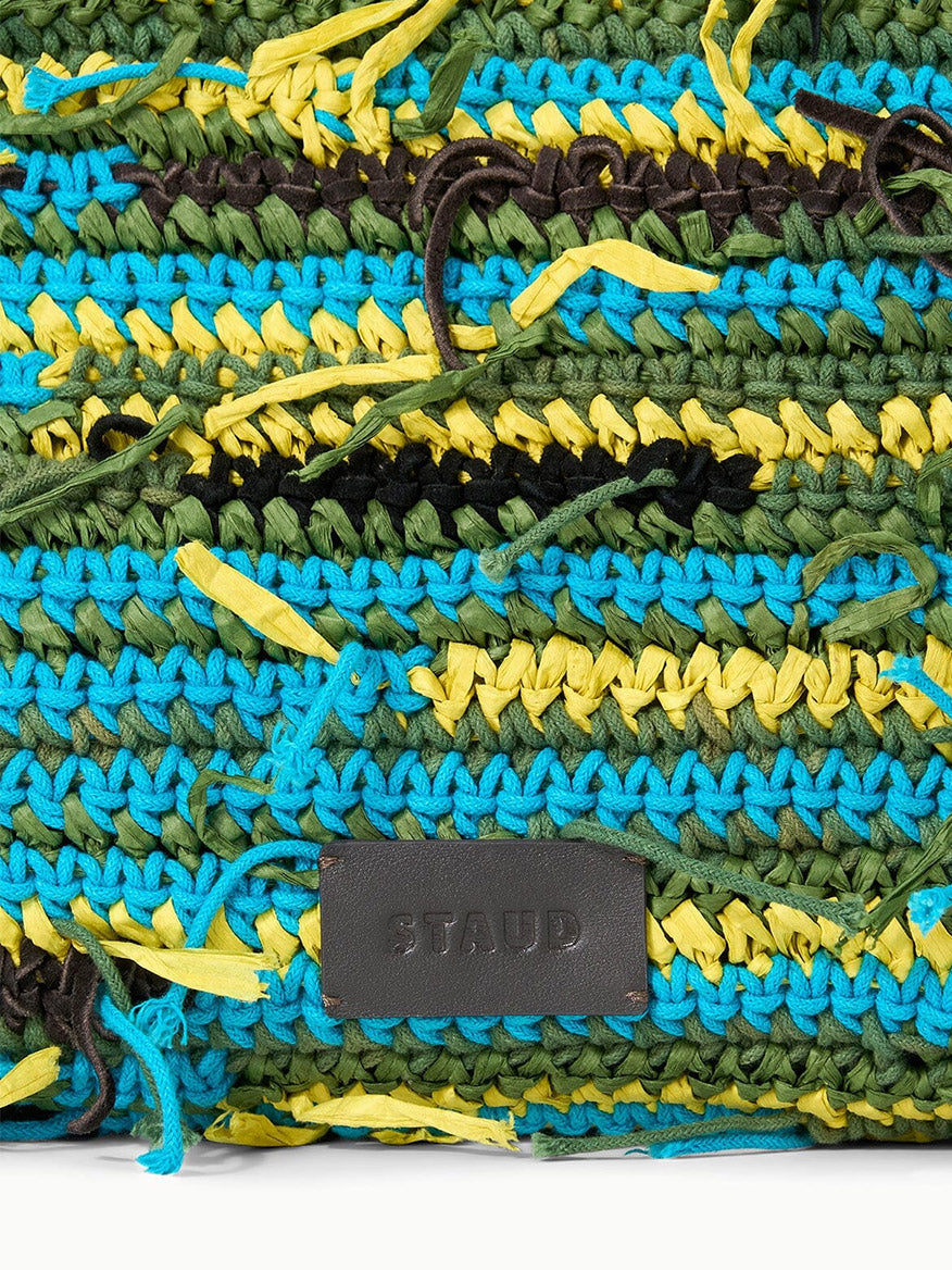 Close-up of the STAUD Christos Large Tote in Happy Sack, featuring multicolored stripes in green, blue, black, and yellow yarns with a black leather patch embossed with "STAUD.