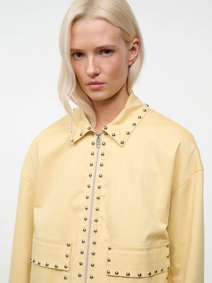 A person with blonde hair wears the STAUD Clarke Jacket in Butter Studs, which features a boxy silhouette and metal studs on the collar, zipper, and pockets, standing against a plain background.