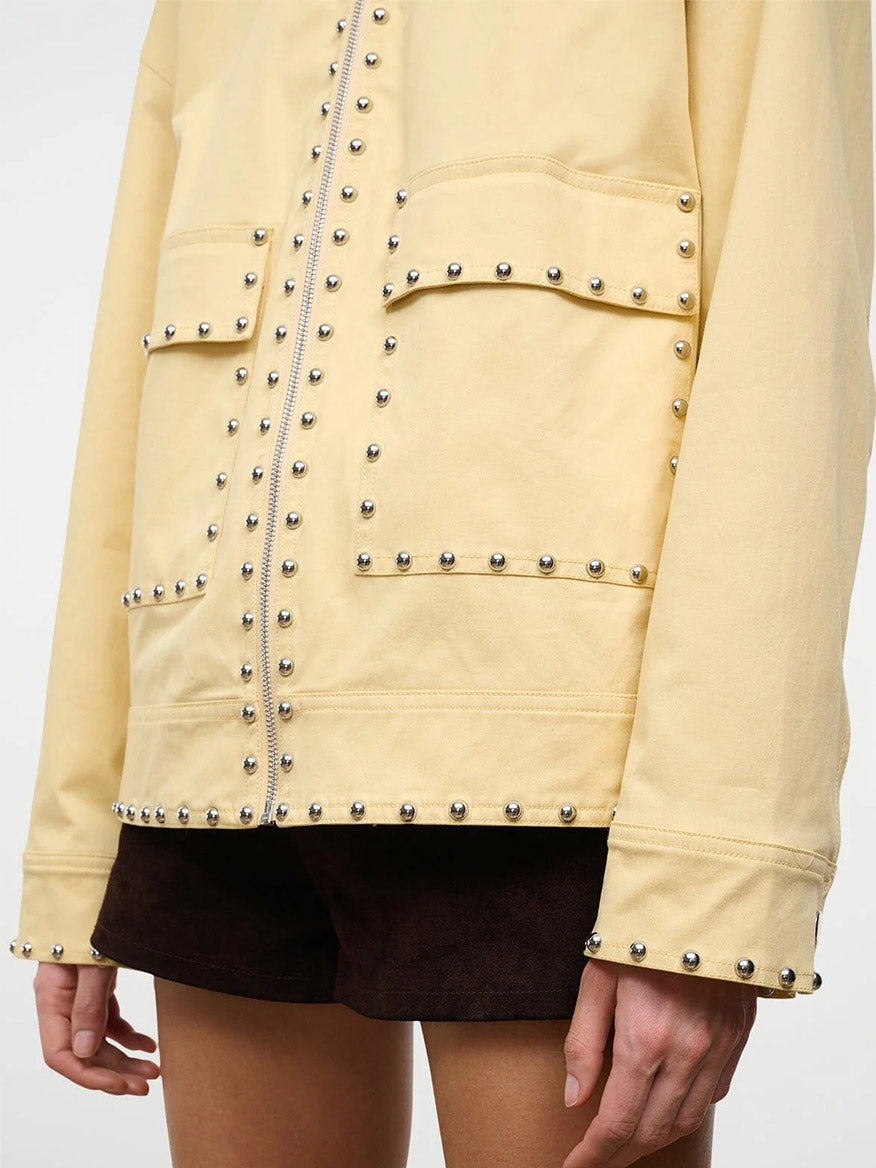 Someone is wearing the STAUD Clarke Jacket in Butter Studs, a light yellow jacket with metal studs, large front pockets, and a boxy silhouette, paired with dark shorts.