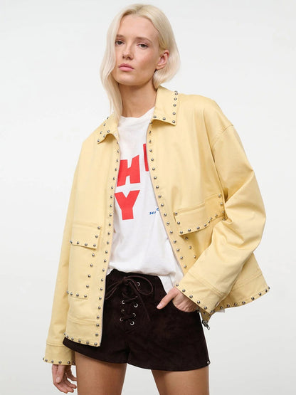 A woman with long blonde hair models the STAUD Clarke Jacket in Butter Studs over a white graphic tee and black lace-up shorts, standing against a plain white background.